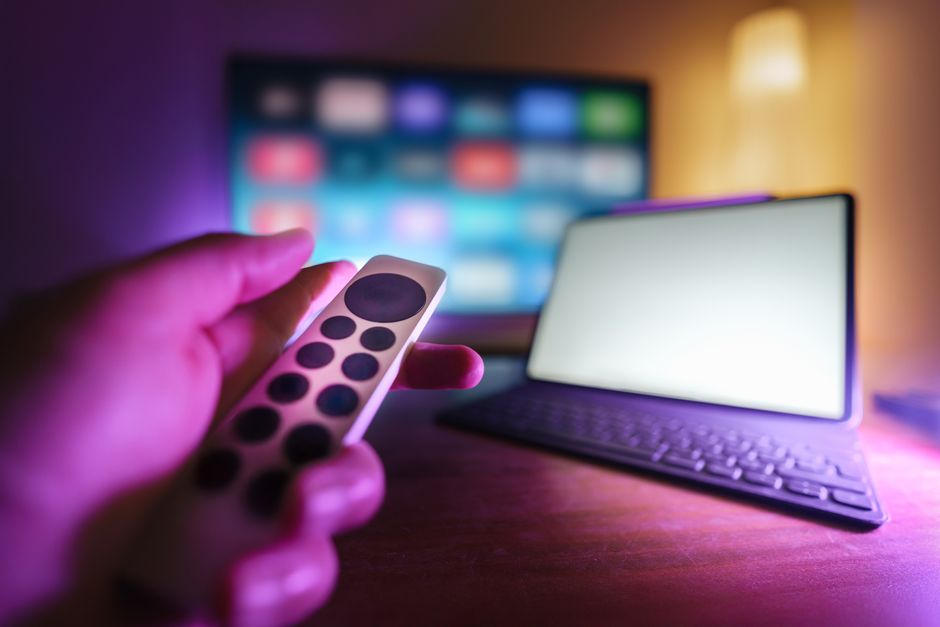 A hand using a remote to control a TV and laptop in a cozy room setting, perfect for relaxation.
